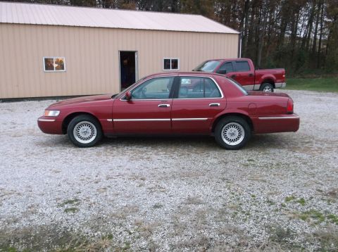 1999 Mercury Grand Marquis - A red 1999 Mercury Grand Marquis is parked on a gravel lot in front of a tan building, with a red pickup truck visible behind it.