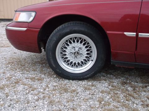 1999 Mercury Grand Marquis - The front wheel and fender of a red 1999 Mercury Grand Marquis are visible, with a mesh alloy rim and chrome trim.