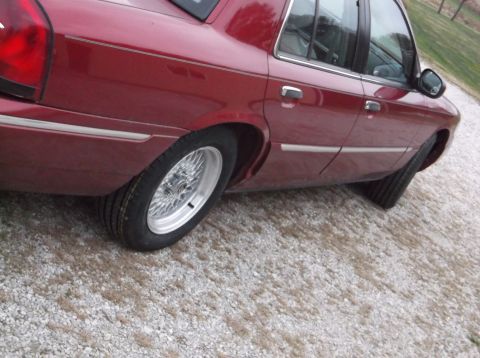 1999 Mercury Grand Marquis - Rear view of a red sedan with chrome trim and wire wheels parked on a gravel surface.