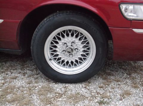 1999 Mercury Grand Marquis - Close-up of a silver lacy-spoke alloy wheel and tire with a Mercury center cap on a maroon sedan over a gravel surface.