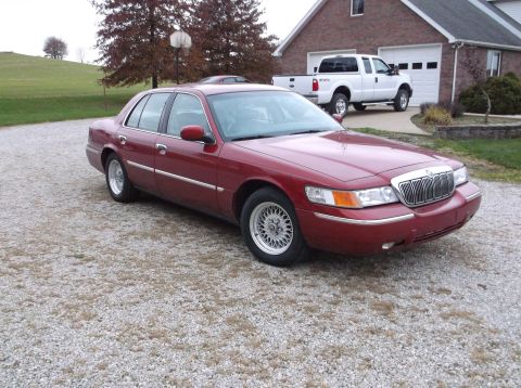 1999 Mercury Grand Marquis - A red sedan is parked on a gravel driveway in front of a brick house with a white pickup truck in the background.