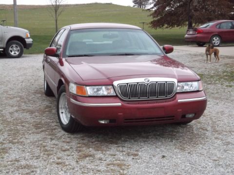 1999 Mercury Grand Marquis - Front view of a red 1999 Mercury Grand Marquis sedan parked on a gravel lot with another vehicle and a dog in the background.