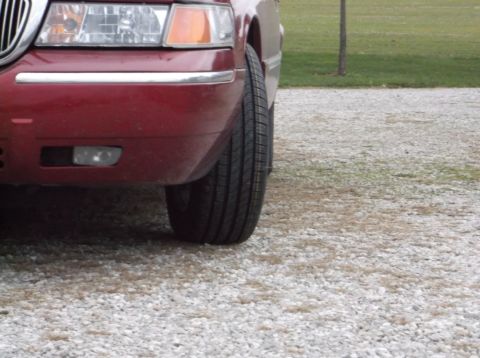 1999 Mercury Grand Marquis - Close-up of the front corner of a red sedan, highlighting the headlight and tire against a gravel surface.