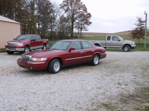 1999 Mercury Grand Marquis - A red 1999 Mercury Grand Marquis is parked on a gravel lot with two pickup trucks in the background.