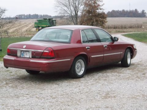 1999 Mercury Grand Marquis - A red sedan with wire wheels is parked on a gravel road with a combine in the background.