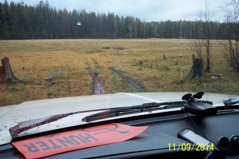 1990 Ford Bronco - View from inside a vehicle as it navigates a muddy track through a field, with a forest bordering the horizon.