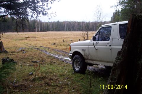 1990 Ford Bronco - A white Ford Bronco is parked off-road on a muddy path, surrounded by a field and forest.