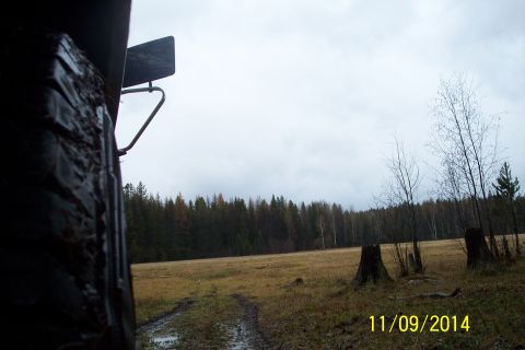 1990 Ford Bronco - A muddy tire and fender are visible in the foreground, with a field and forest stretching out under an overcast sky.