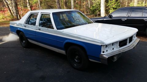 1991 Ford LTD Crown Victoria - A white and blue sedan is parked outside, emitting smoke from the exhaust pipe, with a black car behind it.
