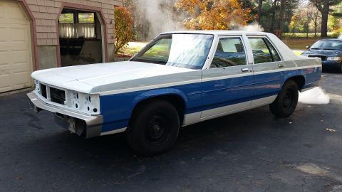 1991 Ford LTD Crown Victoria - A white and blue Ford Crown Victoria is parked in front of a garage, emitting smoke from its exhaust pipe.