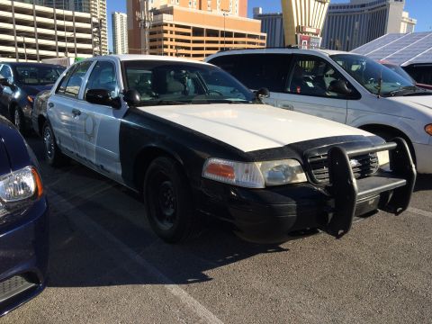 1998 Ford Crown Victoria - A two-tone police car is parked in a lot with other vehicles and buildings in the background.