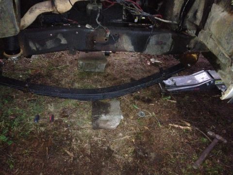 1971 Ford F-250 - Rear leaf-spring pack supported on concrete blocks beneath a truck frame at night, with wiring and a hammer on the ground.