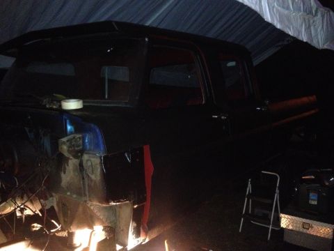 1971 Ford F-250 - A dark pickup truck is seen in profile, partially covered and undergoing bodywork, with visible red accents and exposed metal.