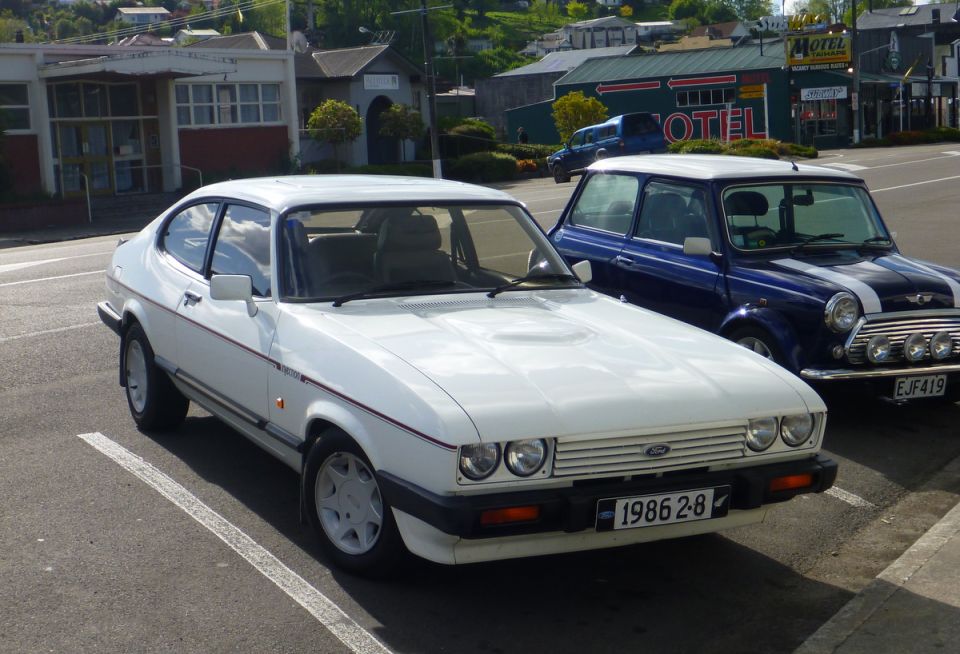 White Ford Capri parked next to a blue Mini Cooper. License plate on Capri reads 1986 2-8
