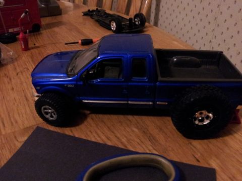 1995 Ford Bronco - A blue pickup truck model sits on a wooden surface, with other toy vehicle parts scattered around.