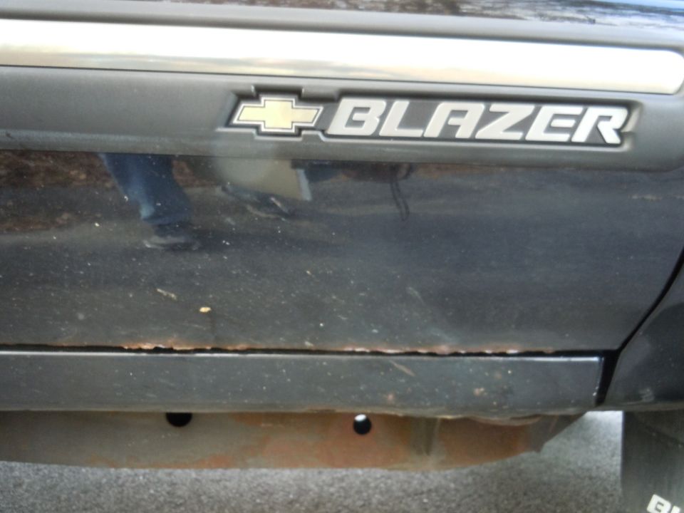 Side view of a dark-colored vehicle showing a 'Blazer' badge with a Chevrolet logo. Rust is visible along the lower body panel