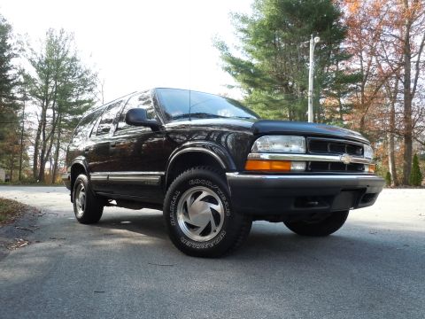 2001 Ford Crown Victoria - Black Chevrolet Blazer with chrome wheels and Goodyear tires sits on a paved road in front of autumn trees.