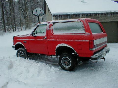 1990 Ford Bronco - A red 1990 Ford Bronco is partially buried in snow, parked in front of a house with icicles hanging from the roof.
