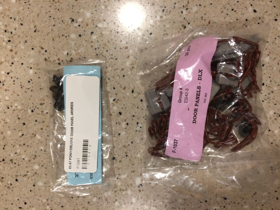 Two bags of car parts. One bag has black clips and a blue card labeled "65-67 PONY/DELUXE DOOR PANEL ARMRES". The other bag has brown clips and gray plastic pieces with a pink label that says "DOOR PANELS - DLX"