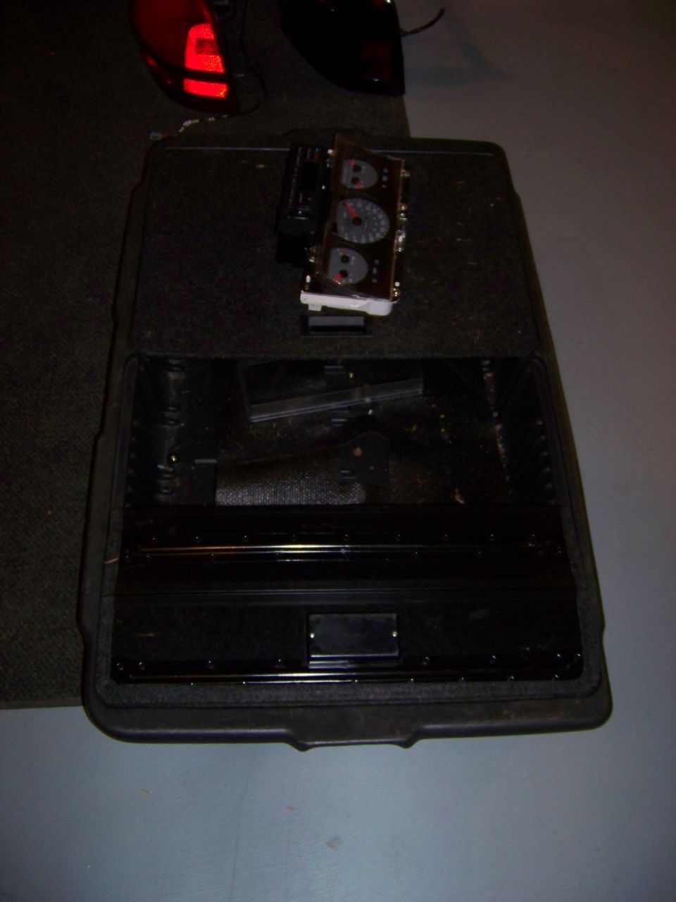 Car dashboard instrument cluster sitting on top of a black storage bin. A red taillight is visible in the background