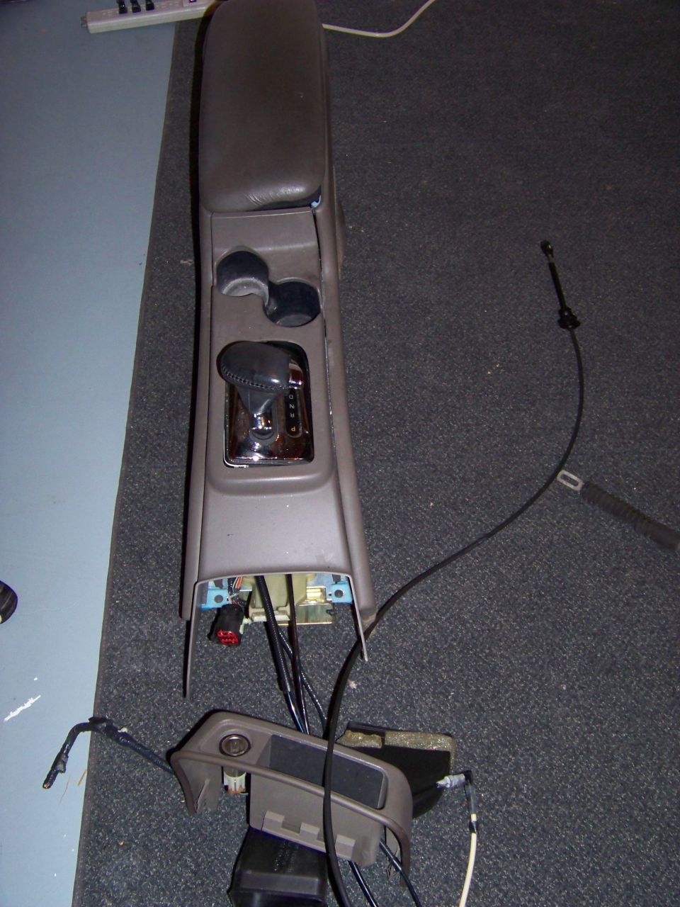 Car's center console and gear shifter removed, showing wiring underneath. A separate trim piece is also visible