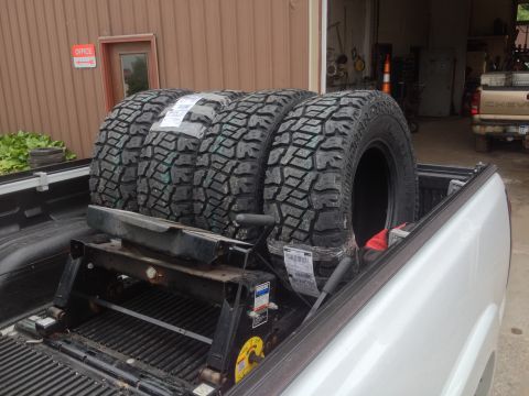 2006 Chevrolet Silverado 2500 - Four large off-road tires are loaded into the bed of a white pickup truck, ready for transport.