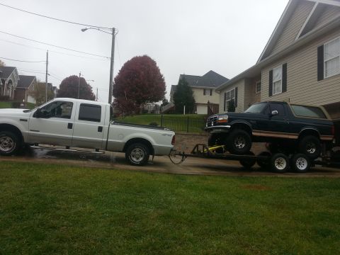 1995 Ford Bronco - A white pickup truck tows a dark green Ford Bronco on a trailer in front of a house.
