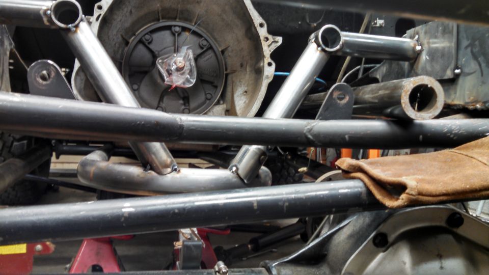 Close-up of a transmission housing with custom metal tubing welded around it. A brown leather glove rests on a metal bar
