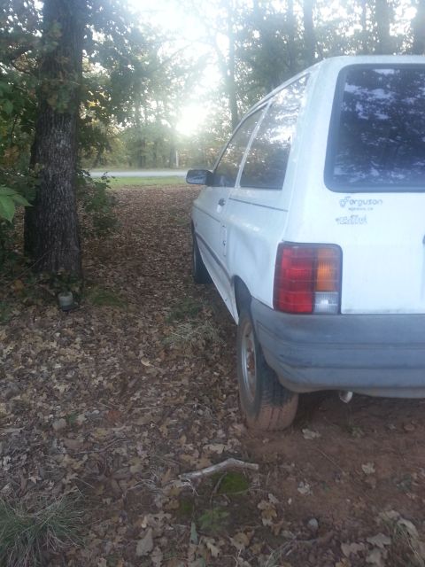 1991 Ford Festiva - Rear view of a white 1991 Ford Festiva parked on a dirt road with trees in the background.