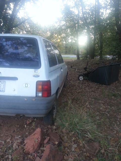 1991 Ford Festiva - The rear of a white 1991 Ford Festiva is visible, parked on uneven ground near trees with a trailer to its right.