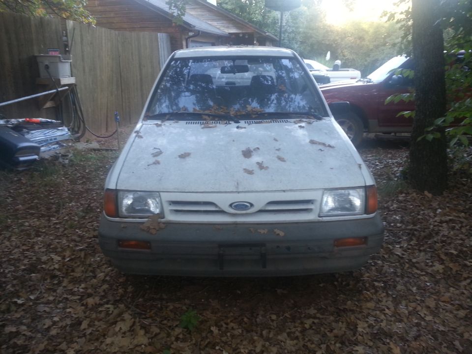 1991 Ford Festiva front view, parked in a yard with leaves on the hood. Another vehicle is visible behind it