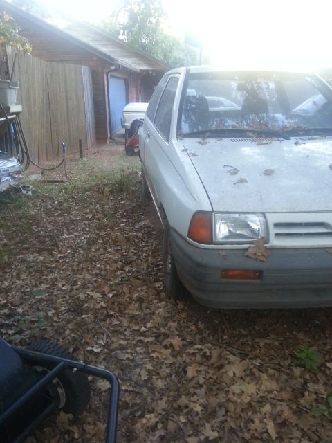 1991 Ford Festiva - A white hatchback is parked in a yard next to a wooden structure, with leaves scattered on the ground.
