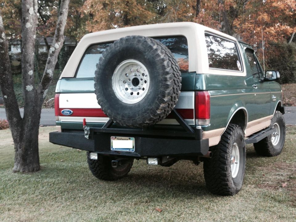 Rear view of a green Ford Bronco with a spare tire mounted on the back. It has a tan top and a custom bumper