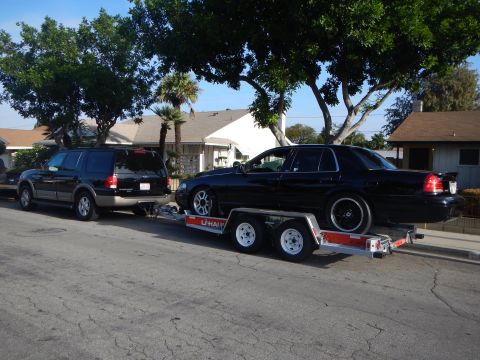 2003 Ford Crown Victoria - black SUV tows a black Ford sedan loaded on a tandem-axle U-Haul car trailer along a tree-lined residential street.