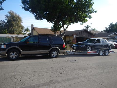 2003 Ford Crown Victoria - Black Ford Expedition with two-tone trim parked curbside towing a black sedan on a U-Haul trailer in a neighborhood.