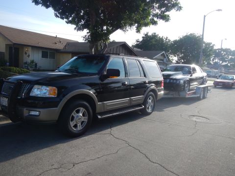 2003 Ford Crown Victoria - Front three-quarter view of a black Ford Expedition towing a black sedan on a U-Haul trailer along a suburban street.