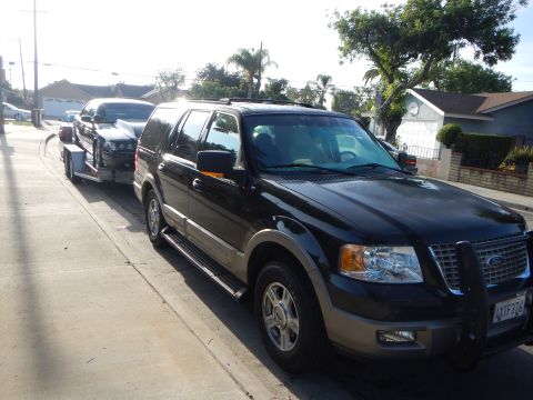 2003 Ford Crown Victoria - Black Ford Expedition with brush guard towing a black sedan on a trailer down a sunlit residential street.
