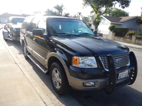 2003 Ford Crown Victoria - Front three-quarter view of a black Ford Expedition with brush guard towing a black sedan on a trailer at a curb.