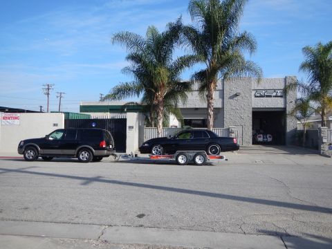 2003 Ford Crown Victoria - black SUV tows a black sedan on a tandem-axle trailer parked along the street outside a gray cinderblock auto shop framed by palm trees.