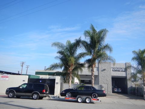 2003 Ford Crown Victoria - black SUV with a trailer carrying a black sedan sits at the curb in front of a gray auto shop under bright blue sky and palms.