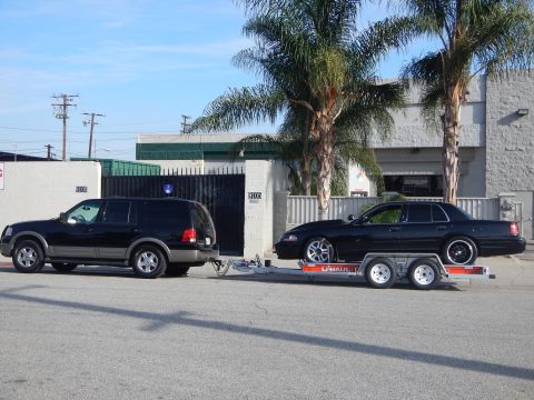 2003 Ford Crown Victoria - black sedan with polished chrome five-spoke wheels rides on a U-Haul flatbed trailer pulled by a black SUV in front of an auto shop.