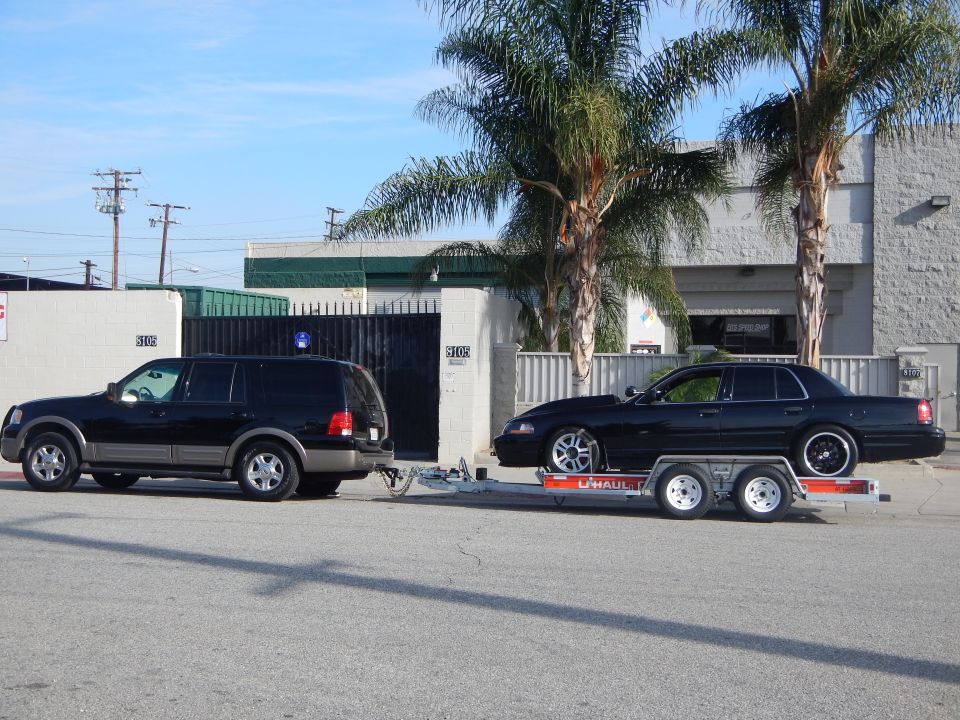 Black sedan with chrome wheels loaded on a U-Haul trailer towed by a black SUV