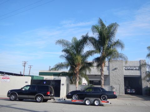 2003 Ford Crown Victoria - black SUV tows a black sedan on a tandem trailer along the street outside a gray auto shop shaded by large palm trees.