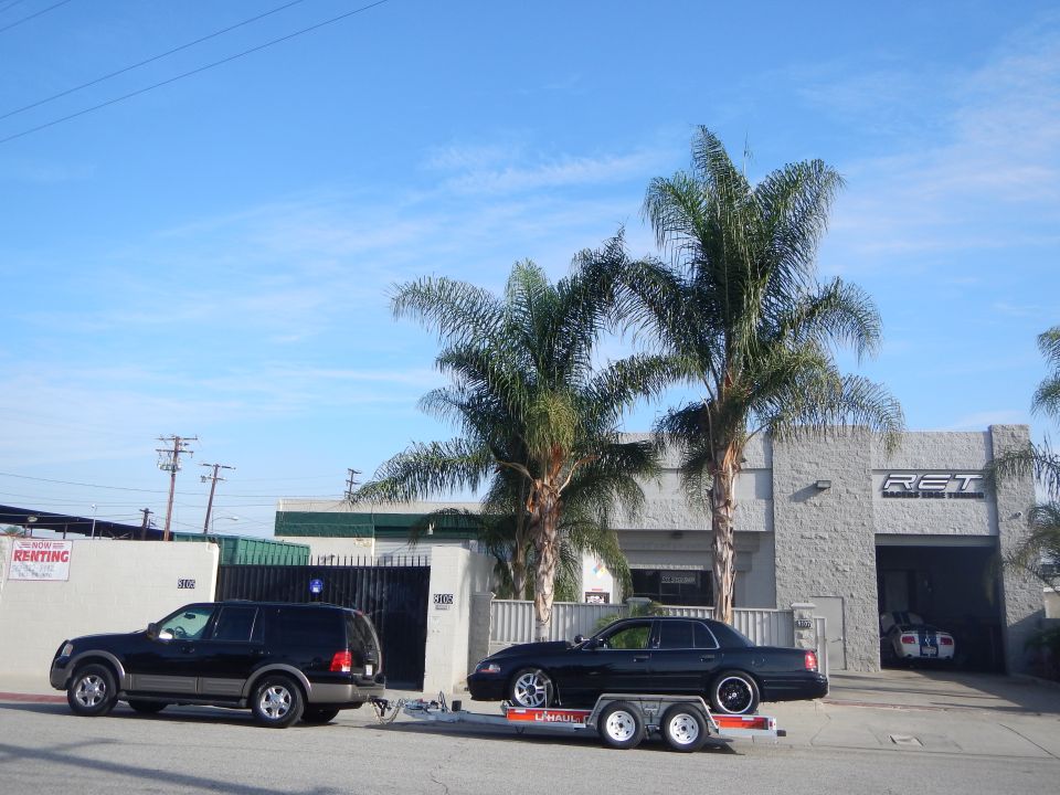 Black SUV towing a black sedan on a trailer past a gray shop building with palm trees