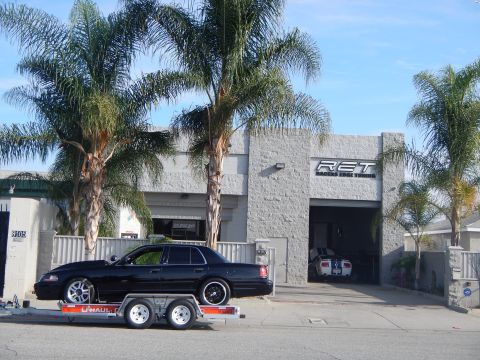 2003 Ford Crown Victoria - lowered black sedan on chrome wheels sits loaded on a U-Haul trailer in front of an open gray auto-shop garage bay.