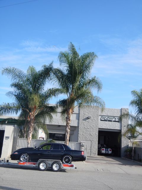 2003 Ford Crown Victoria - black sedan rests on a U-Haul flatbed trailer beneath towering palm trees in front of a gray cinderblock auto shop.
