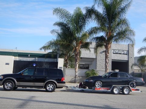 2003 Ford Crown Victoria - black SUV with a trailer holding a black sedan is parked at the curb outside a gray auto shop ringed by palm trees.