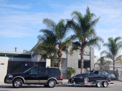 2003 Ford Crown Victoria - black SUV tows a black sedan on a tandem trailer parked in front of a gray auto shop framed by tall palm trees.