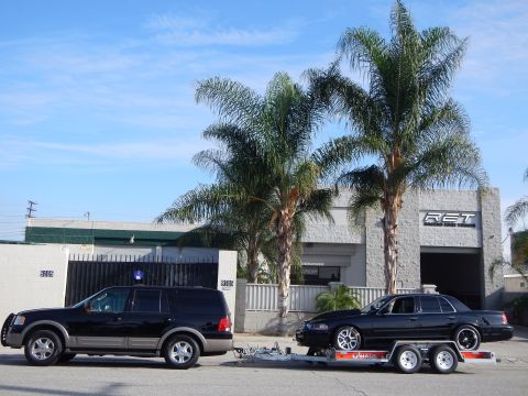2003 Ford Crown Victoria - black SUV with a U-Haul trailer carrying a black sedan sits curbside before a gray auto shop under blue sky and palms.