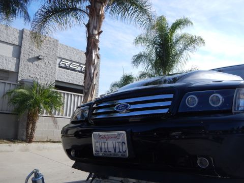 2003 Ford Crown Victoria - Close-up of a black Ford sedan's chrome-slat grille with halo projector headlights and a California EVIL VIC plate, loaded on a trailer.